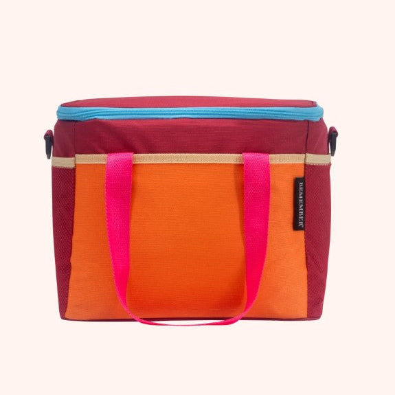 A rectangular cooler bag with a color-blocked design featuring orange, magenta, and dark red panels, with a bright pink handle and a light blue zipper trim, shown against a solid pale pink background.