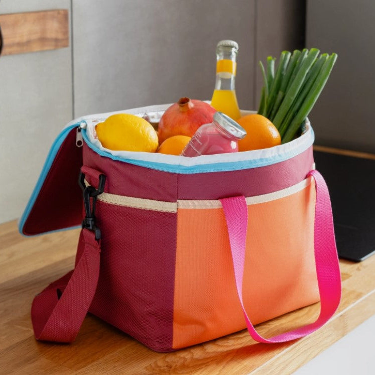 A reusable insulated cooler bag with a maroon and orange color-blocked design, pink handles, and a light blue zipper trim, shown open on a wooden countertop filled with fruit and a drink.