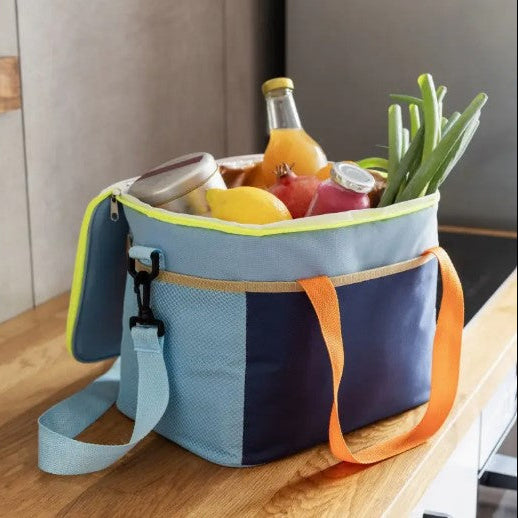 A light blue and navy insulated lunch bag with orange straps, filled with groceries including a bottle of juice, a lemon, and green onions, sitting on a wooden table.