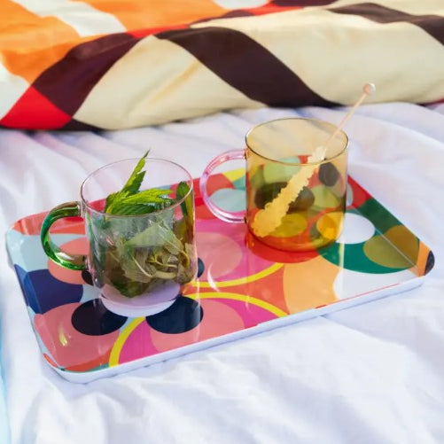 A colorful patterned tray holding two translucent mugs on a bed. One clear mug has mint leaves and a green handle, and the other amber-tinted mug has a yellow drink and a straw.