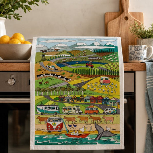A kitchen tea towel with a colorful illustrated landscape scene featuring campers, cows, houses, mountains, and a whale, hanging on an oven handle in a kitchen setting.