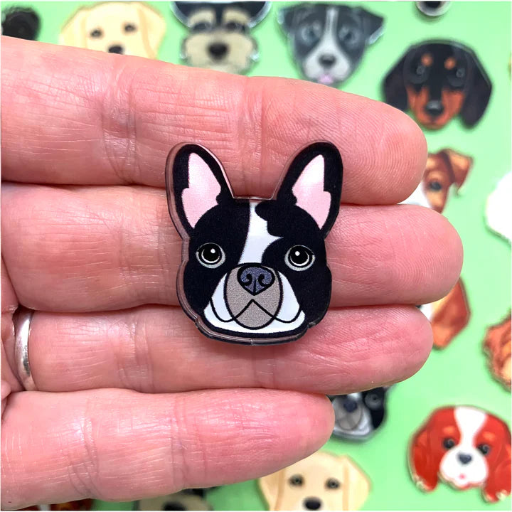 French bulldog shaped pin held between fingers with a colorful background of dog illustrations.