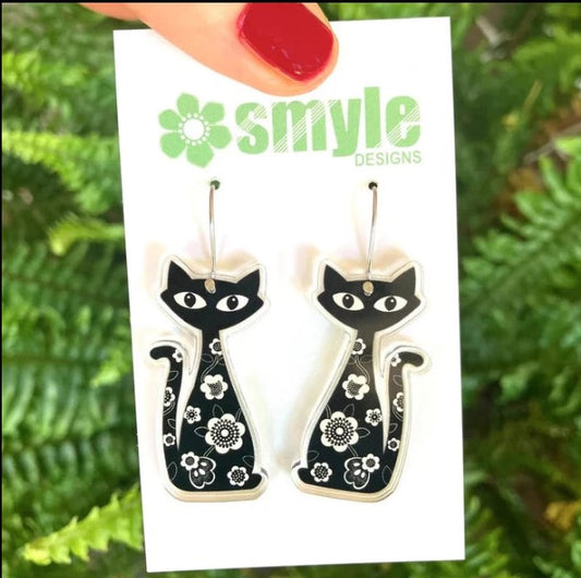 Cat-shaped earrings on a Smyle Designs card with a green leafy background